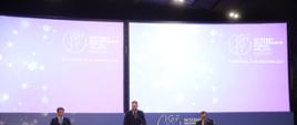 Szczyt Cyfrowy ONZ – IGF 2021. Na scenie przy mównicach stoją trzej mężczyźni: Krzysztof Szubert, minister Janusz Cieszyński i Liu Zhenmin. Fot. PAP