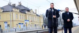 Minister Czarnek stoi na peronie kolejowej stacji i mówi do mikrofonu na stojaku, za nim stoi mężczyzna w czarnej kurtce, w głębi żółty stary budynek stacji.