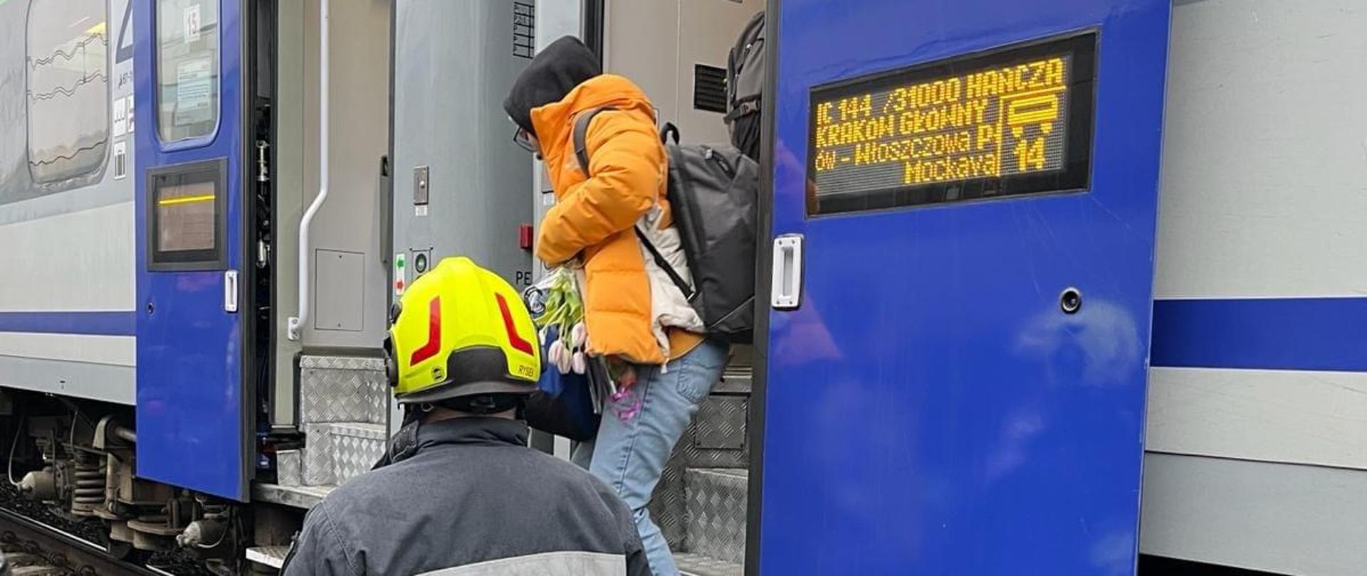 Strażak OSP pomagający pasażerowi w bezpieczny sposób przejść do sprawnego pociągu podstawionego przez przewoźnika.
