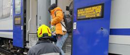 Strażak OSP pomagający pasażerowi w bezpieczny sposób przejść do sprawnego pociągu podstawionego przez przewoźnika.