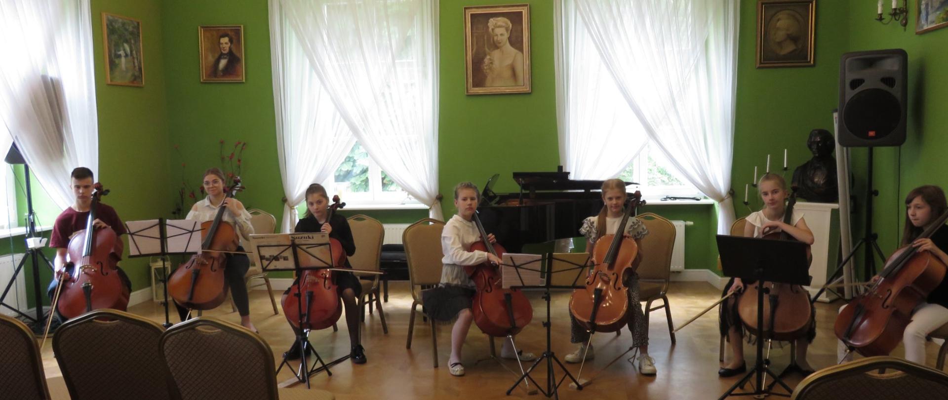 Zespół wiolonczelowy siedzi na krzesłach, 6 dziewczynek i 1 chłopiec, trzymają instrumenty, w ręku smyczki, w tle fortepian i portret Melanii Bronikowskiej i Fryderyka Chopina
