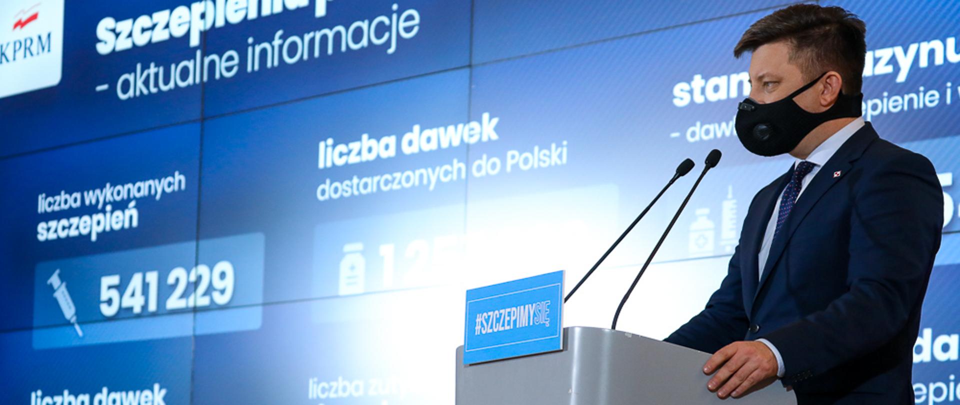 Szef KPRM Michał Dworczyk podczas konferencji prasowej. W tle ekran z informacjami nt. szczepień przeciwko Covid-19.