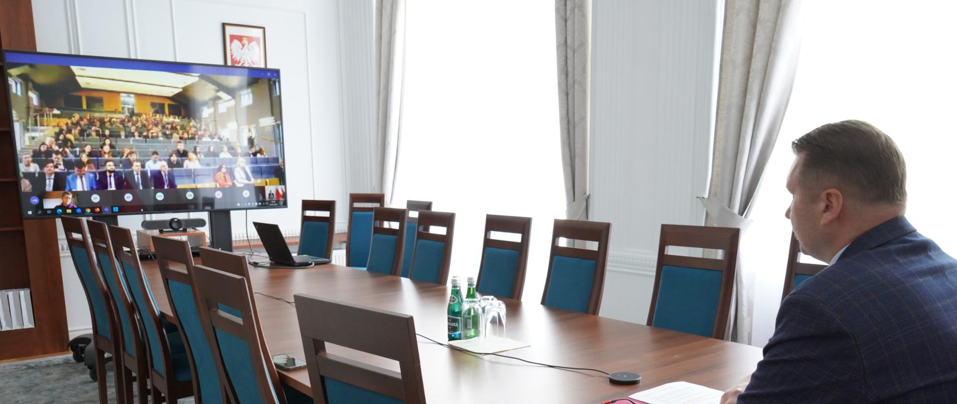 Stół wokół niego poustawiane krzesła, po jednej stronei ekran z widokiem na aulę uniersytecką, po drugiej siedzą przy stole minister Czarnek. 
