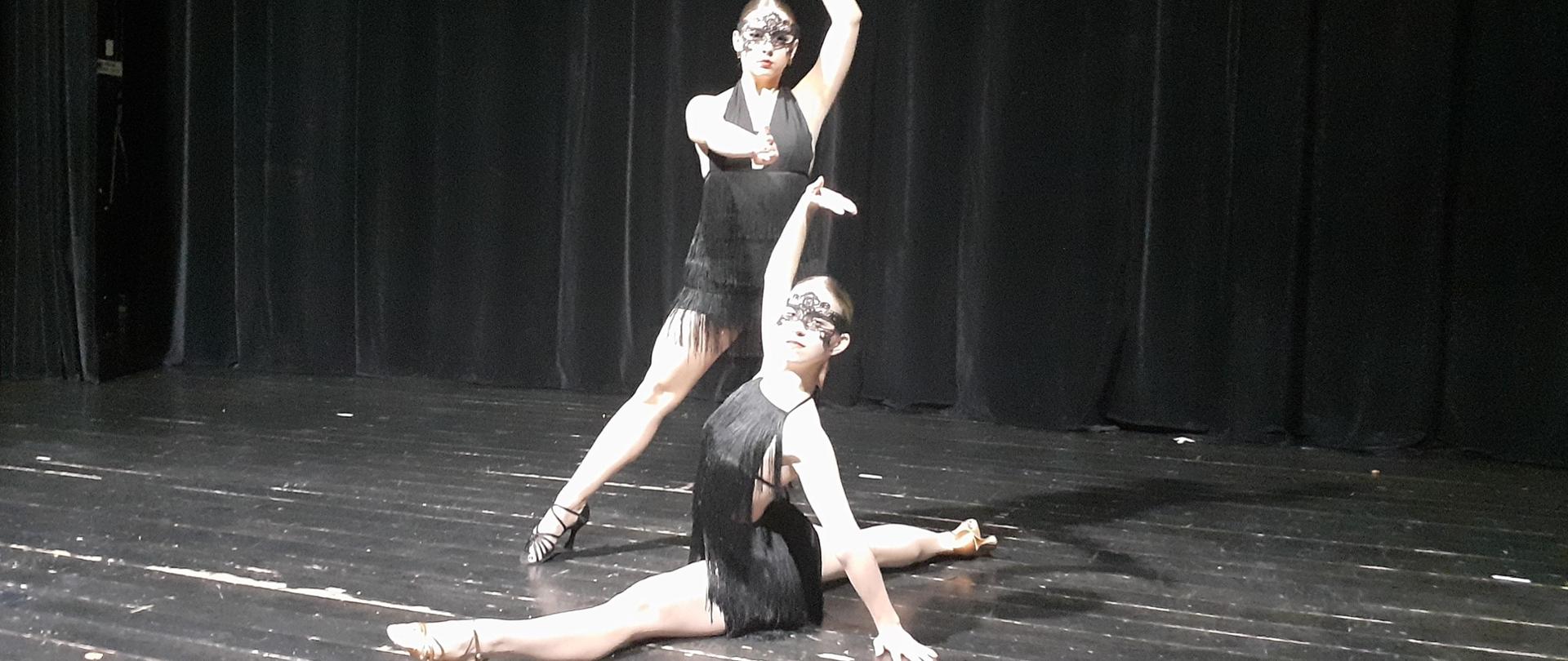 Na zdjęciu dwie uczennice tańczące na scenie teatru. Jedna uczennica robi szpagat, druga za nią tańczy.