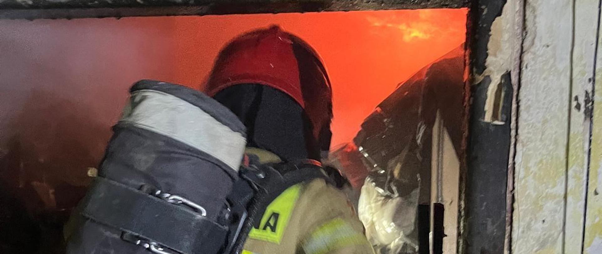 Strażak pracuje wewnątrz spalonego pomieszczenia. Przed nim widać płomienie ognia. 
