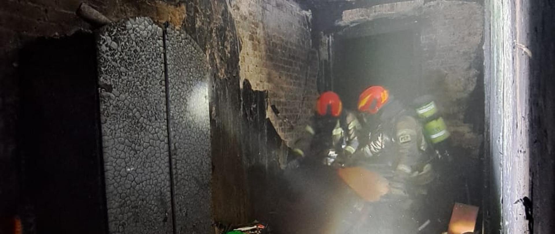 Na zdjęciu widoczni strażacy przeszukujący pomieszczenie w którym doszło do pożaru, na pierwszym planie widoczna najaśnica akumulatorowa i spalona szafa.