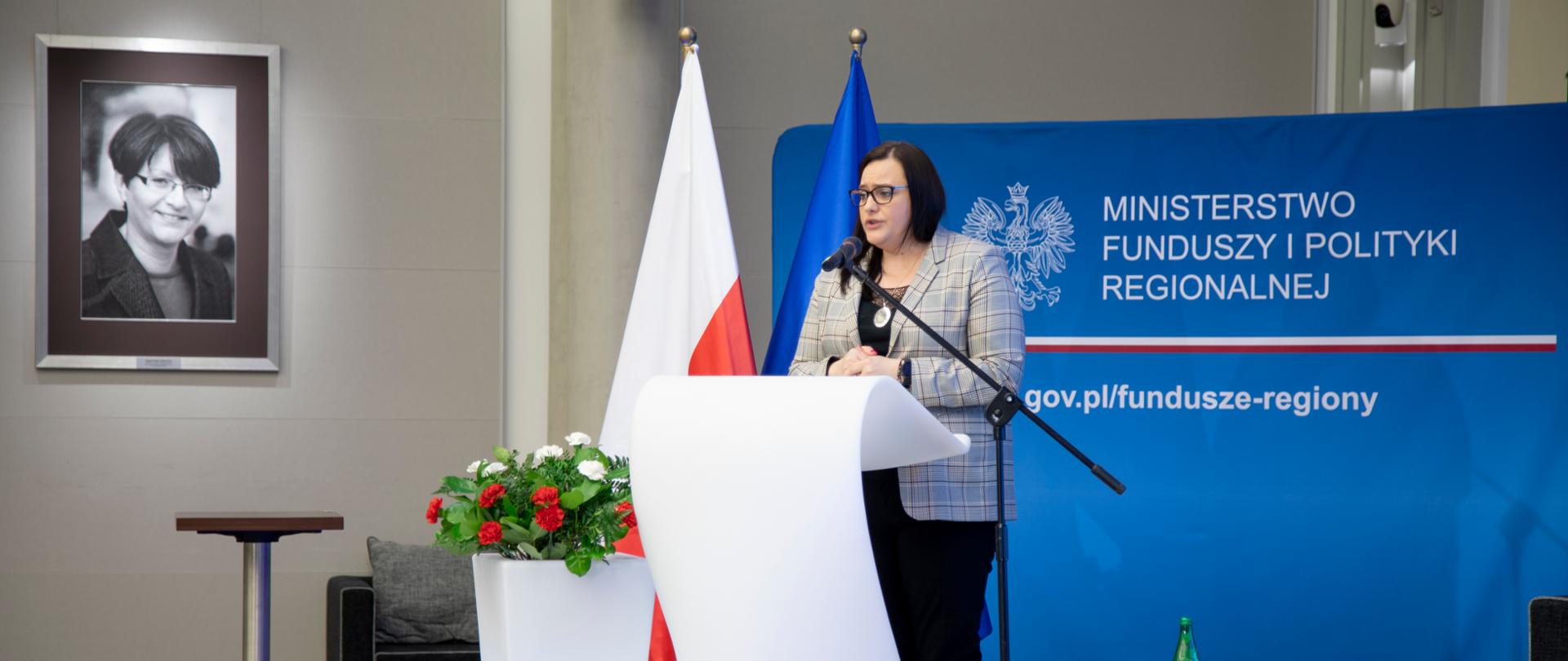 wiceminister Małgorzata Jarosińska-Jedynak mówi do mikrofonu, za nią zdjęcie minister Grażyny Gęsickiej