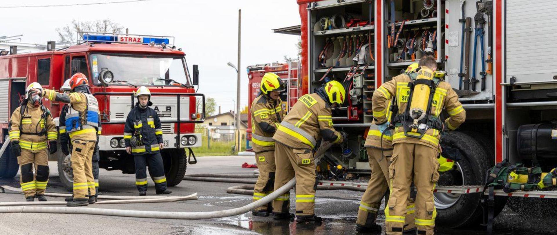Strażacy jednostek ochotniczych straży pożarnych prowadzą działania przy pojazdach pożarniczych. Wśród strażaków jest jeden z Państwowej Straży Pożarnej. Strażacy w umundurowaniu specjalnym z hełmami na głowach.
