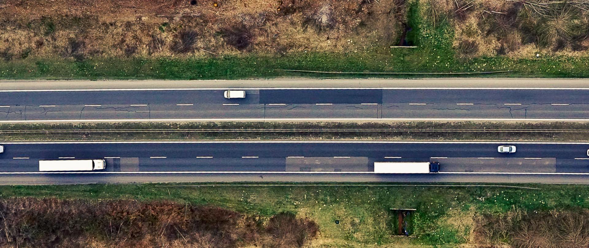 Zdjęcie lotnicze dwujezdniowej drogi krajowej. Na jezdni widocznej samochody osobowe i ciężarowe poruszające się po drodze. Drogę pod kątem prostym przecina niewielki ciek. 