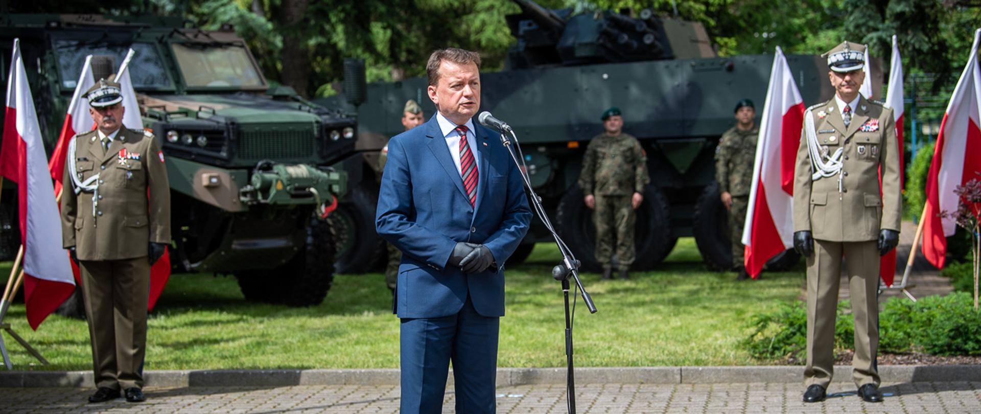 Minister Mariusz Błaszczak przy mikrofonie podczas przemówienia okolicznościowego. W tle generałowie, żołnierze oraz sprzęt wojskowy. Po prawej i lewej stronie flagi Polski.