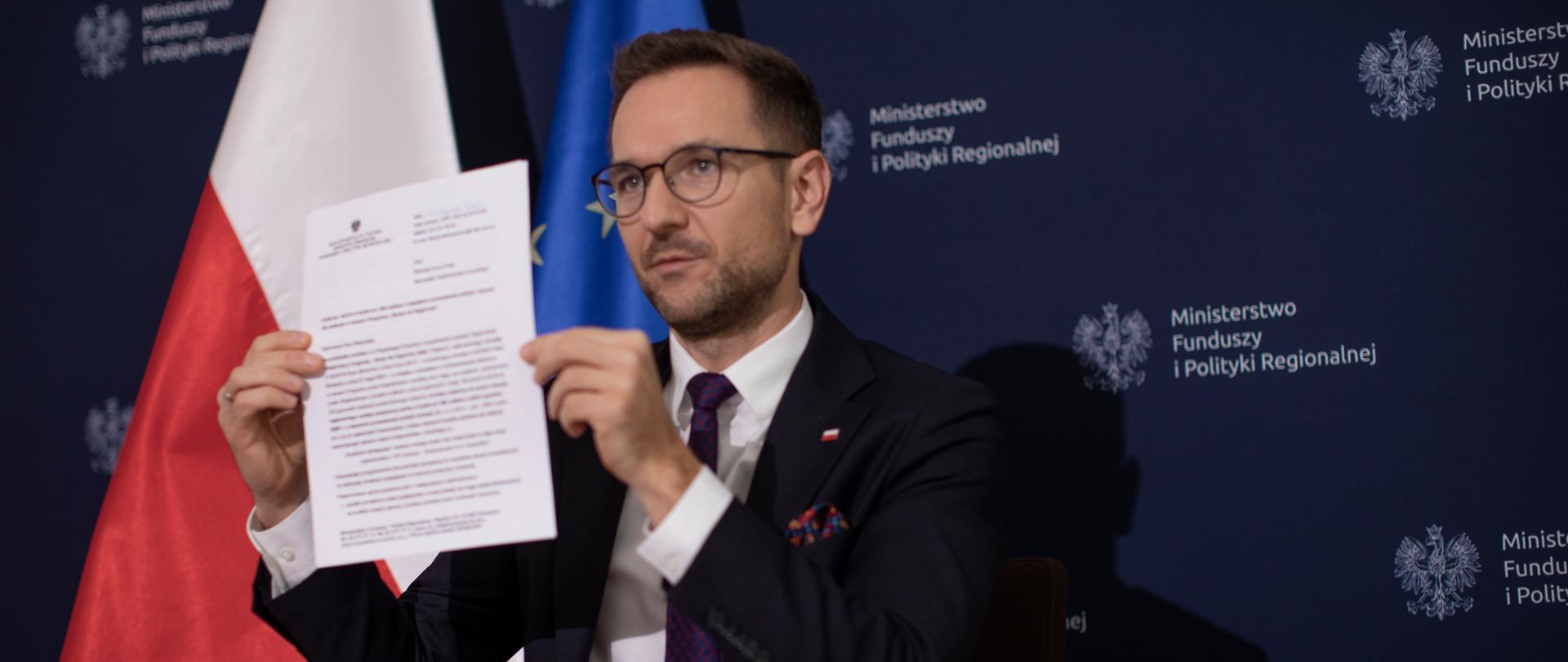 Wiceminister Waldemar Buda siedzi przy stole i prezentuje dokument. Za nim stoją: flagi polski i UE oraz ścianka MFiPR.