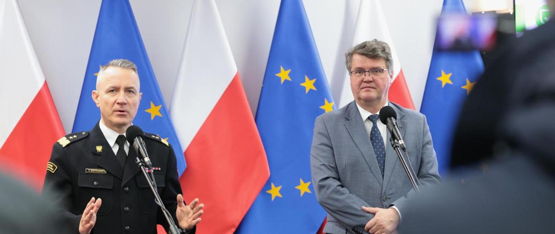 Na zdjęciu widzimy dwóch mężczyzn stojących przed mikrofonami. Mężczyzna z lewej strony kadru to strażak ubrany w mundur galowy koloru czarnego, przemawia, ręce lekko uniesione. Mężczyzna po prawej stronie kadru to cywil ubrany w marynarkę koloru szarego, okulary na oczach. Za mężczyznami widzimy flagi Polski oraz Unii Europejskiej. 