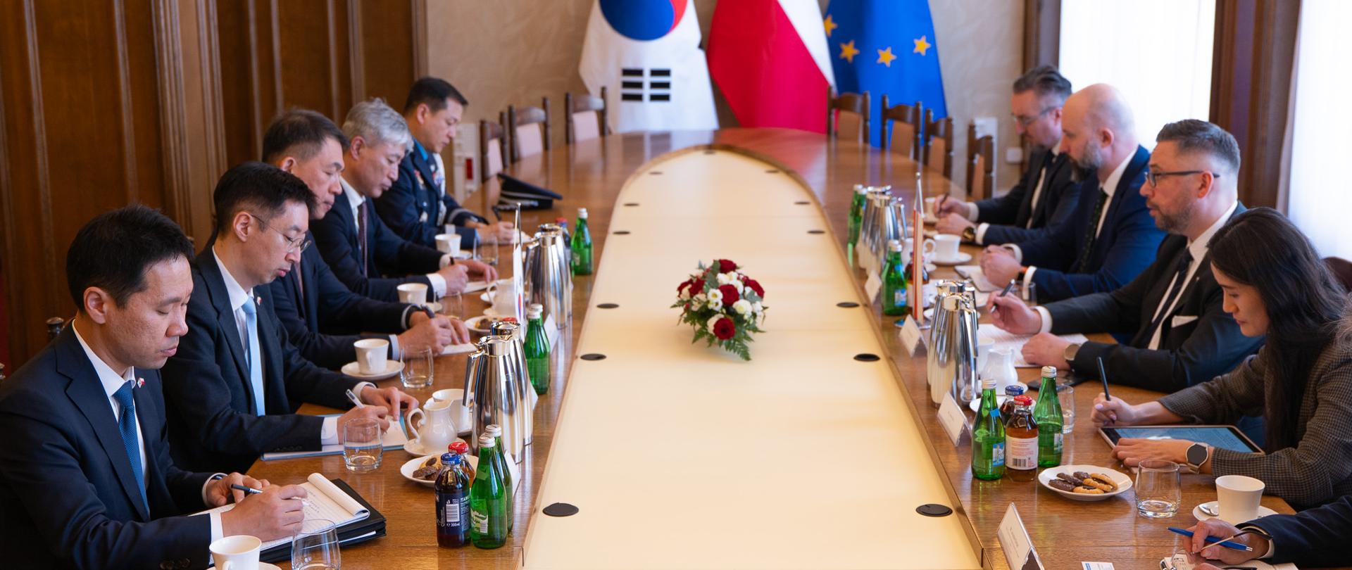 Przedstawiciele delegacji polskiej i koreańskiej siedzą wokół okrągłego stołu. Na stole kwiaty, dzbanki, butelki, szklanki. W tle flagi - południowokoreańska, polska i UE.