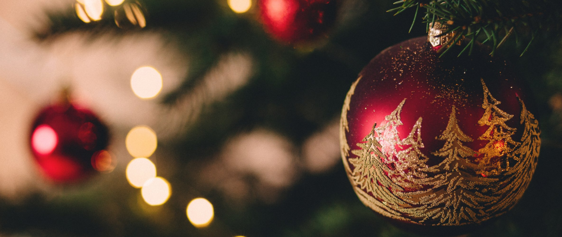 Ilustracja przedstawia czerwoną bombkę ze złotymi choinkami wiszącą na świątecznym drzewku.