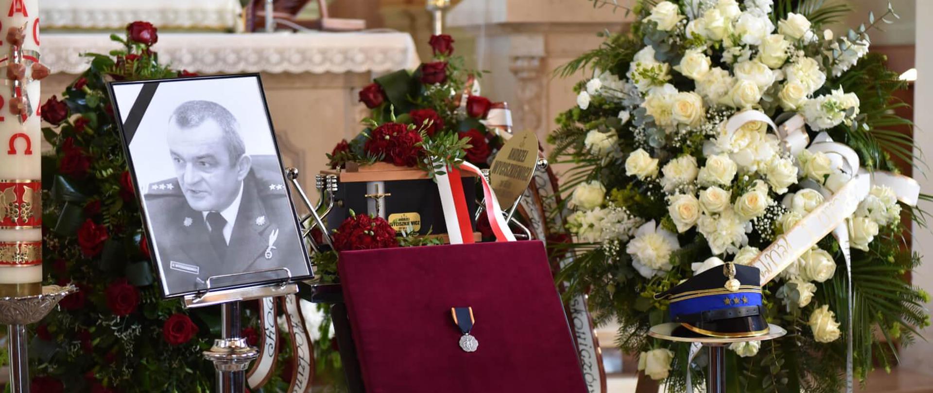 Widok urny z prochami zmarłego, fotografia zmarłego z czarną wstążką, czapka wyjściowa oficera oraz na czerwonej poduszce Krzyż Świętego Floriana. Obok urny z prochami stoją dwa wieńce.