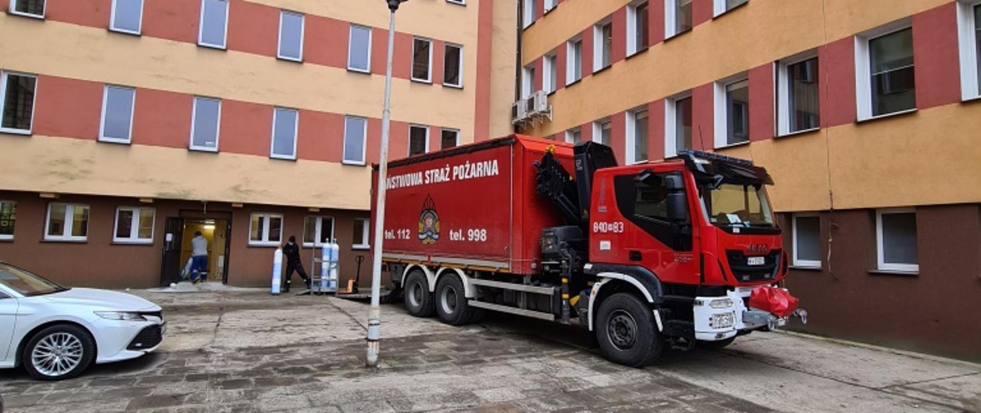 Zdjęcie przedstawia pojazd ciężarowy w kolorze czerwonym z białymi elementami zabudowy, ustawiony przy budynku szpitala powiatowego w Kozienicach. Pojazdem tym zostały dostarczone butle z tlenem medycznym. 