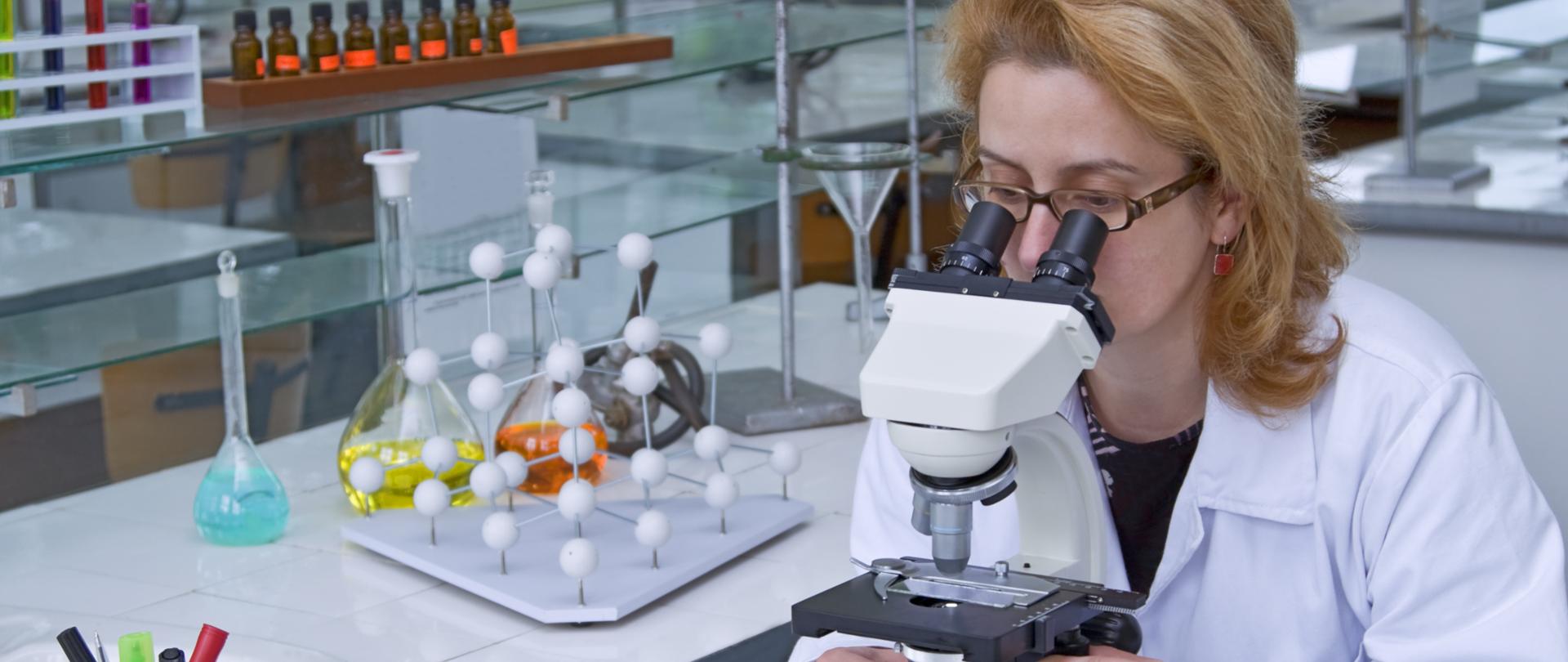 Kobieta w białym fartuchu patrzy przez mikroskop.