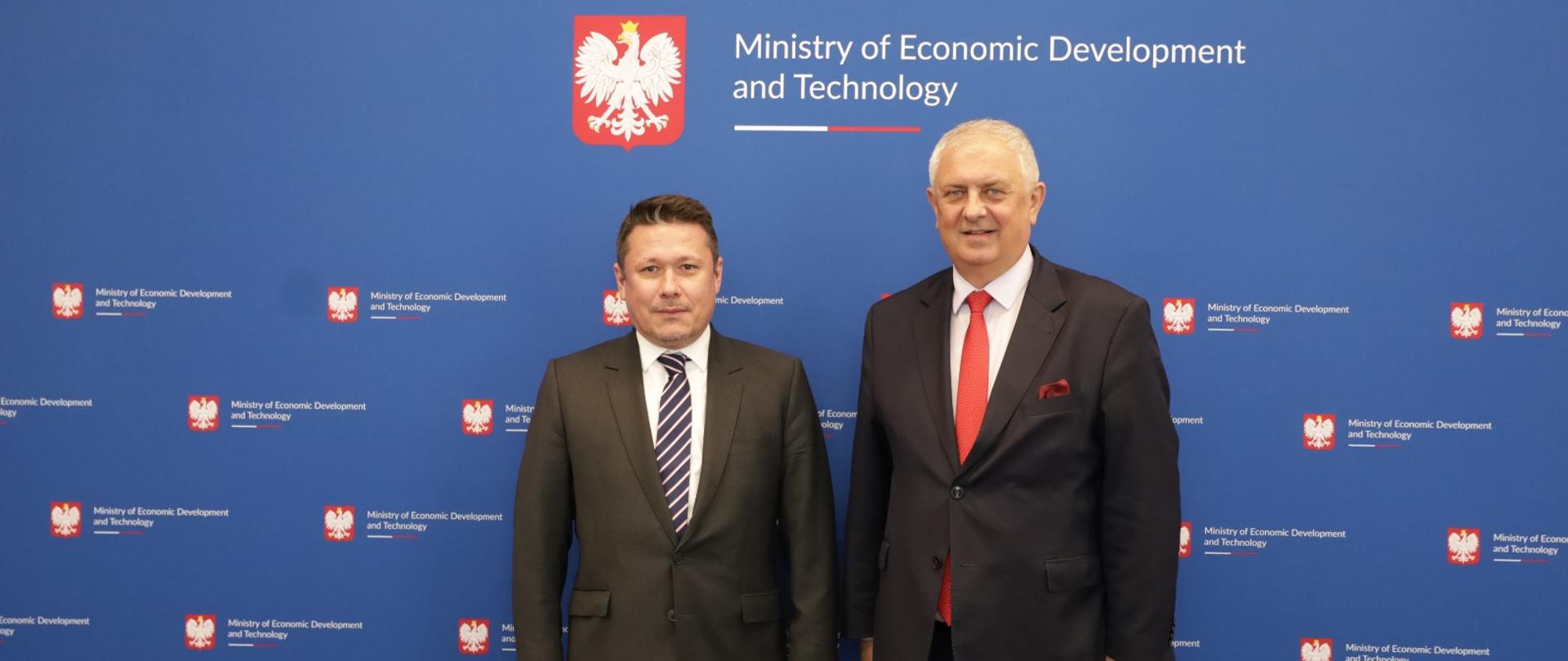 Wiceminister Grzegorz Piechowiak z Ambasadorem Rumunii Cosminem Onisii. Na zdjęciu widać wiceminiser wraz z Ambasadorem Rumunii na granatowym tle z logotypem Ministerstwa Rozwoju i Technologii.