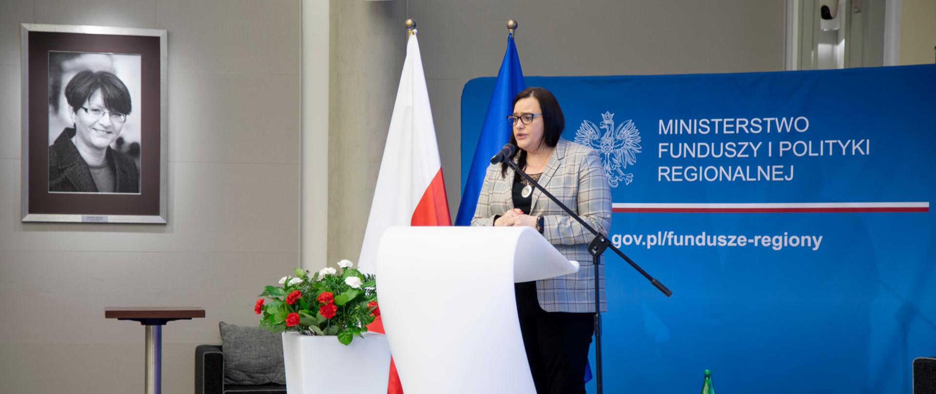 Wiceminister Małgorzata Jarosińska-Jedynak stoi na scenie, za mównicą, mówi do mikrofonu, za nią ścianka z logo MFiPR