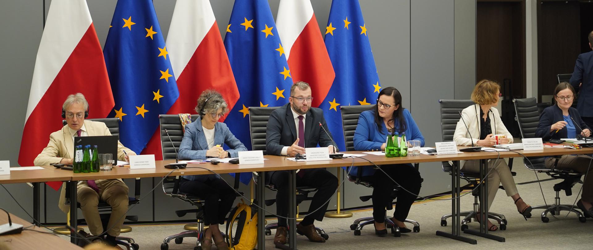 Grupa osób siedzi przy długim stole w sali konferencyjnej. Za nimi flagi PL i UE.
