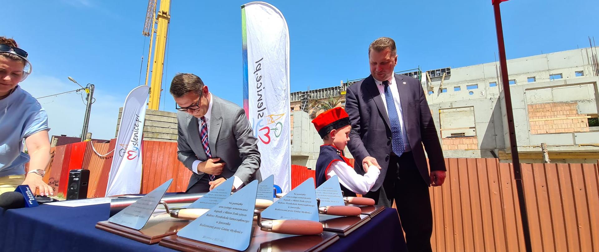 Mały chłopiec w ludowym stroju stoi z ministrem Czarnkiem i ściska mu rękę, obok stół na którym leżą pamiątkowe kielnie z wygrawerowanymi napisami.
