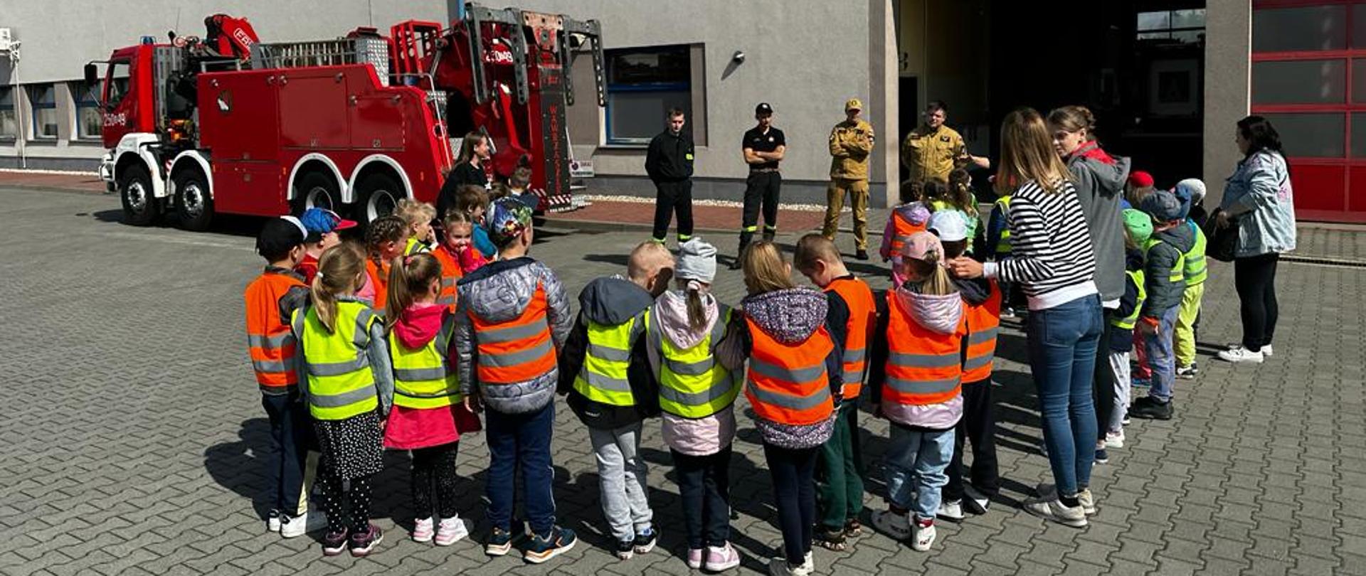 Widok z tyłu na grupę dzieci przedszkolnych wraz z wychowawcami stojącymi przed budynkiem Jednostki Ratowniczo-Gaśniczej CS PSP w głębi wóz bojowy obok którego stoją strażacy