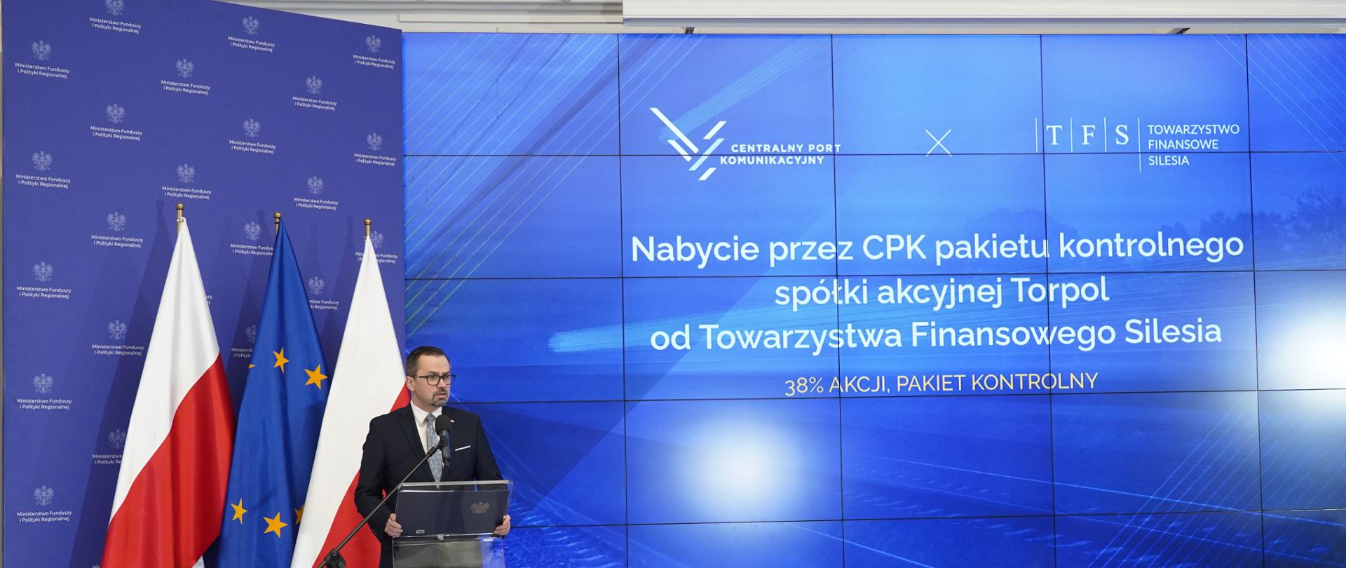 Wiceminister Marcin Horała stoi na scenie z lewej strony, przy pulpicie i mówi do mikrofonu.