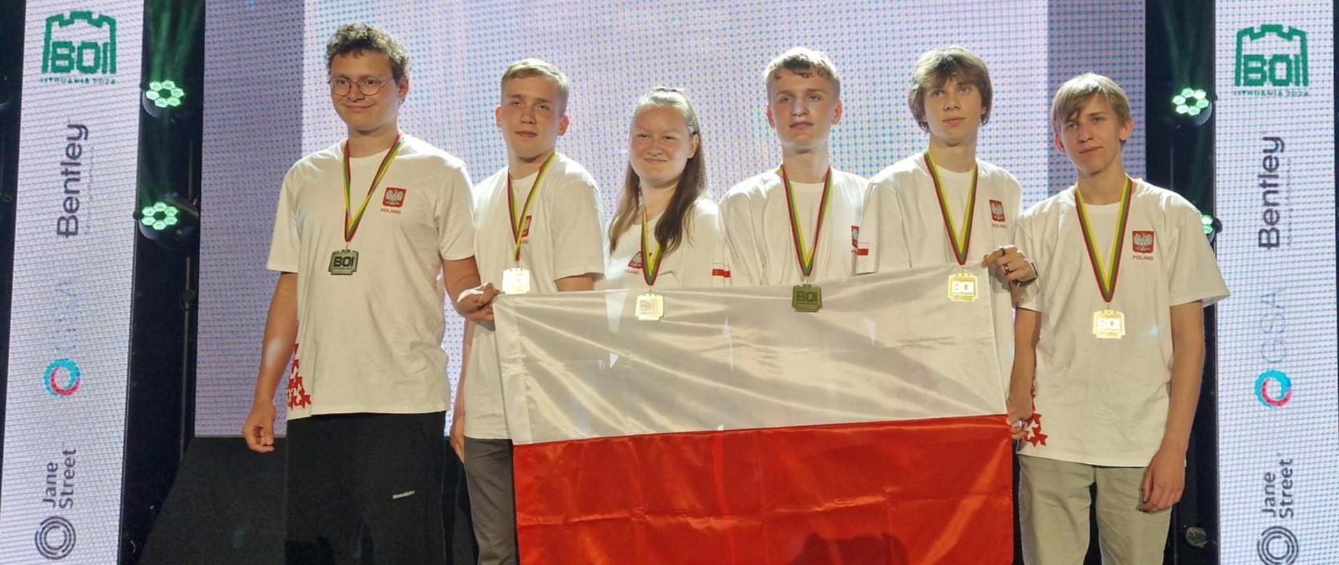 Sześcioro młodych ludzi Ubranych w białe koszuli z godłem Polski stoi na scenie i trzyma biało-czerwoną flagę.