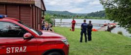 Kolorowa fotografia wykonana na zewnątrz w pochmurny dzień. Przedstawia strażaka ubranego w ubranie do pracy w wodzie koloru czerwonego mówiącego o bezpieczeństwie nad wodą oraz grupę dzieci którzy go słuchają. W pobliżu dwóch policjantów fragment samochodu pożarniczego operacyjnego.