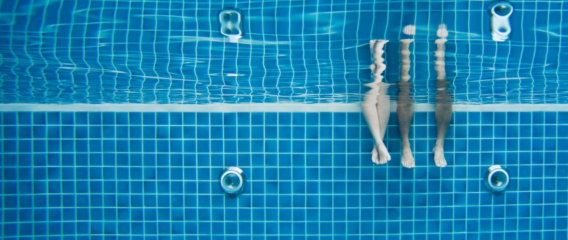 Zdjęcie wykonane pod wodą w basenie. Widać na nim zanurzone nogi dwóch osób. Niebieskie kwadratowe kafelki z białymi fugami. Na ścianie widoczne dwie lampy basenowe.