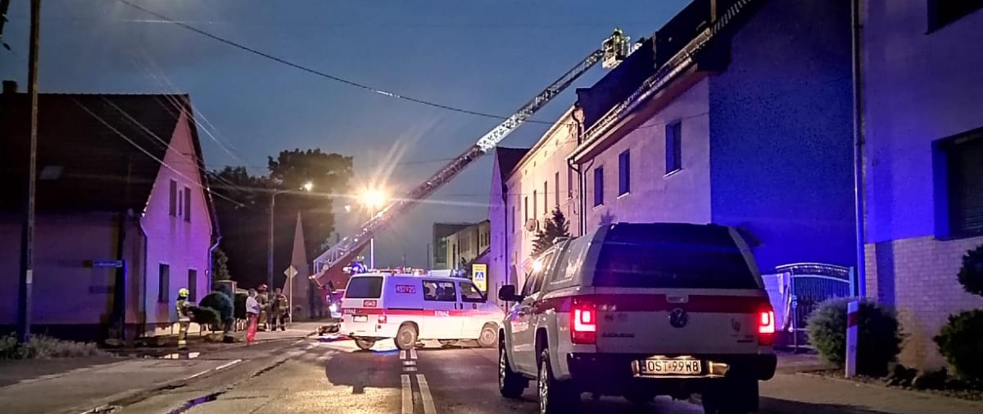 Zdjęcie przedstawia dwa samochody pożarnicze oraz drabinę pożarniczą przy zabezpieczaniu uszkodzonego dachu.