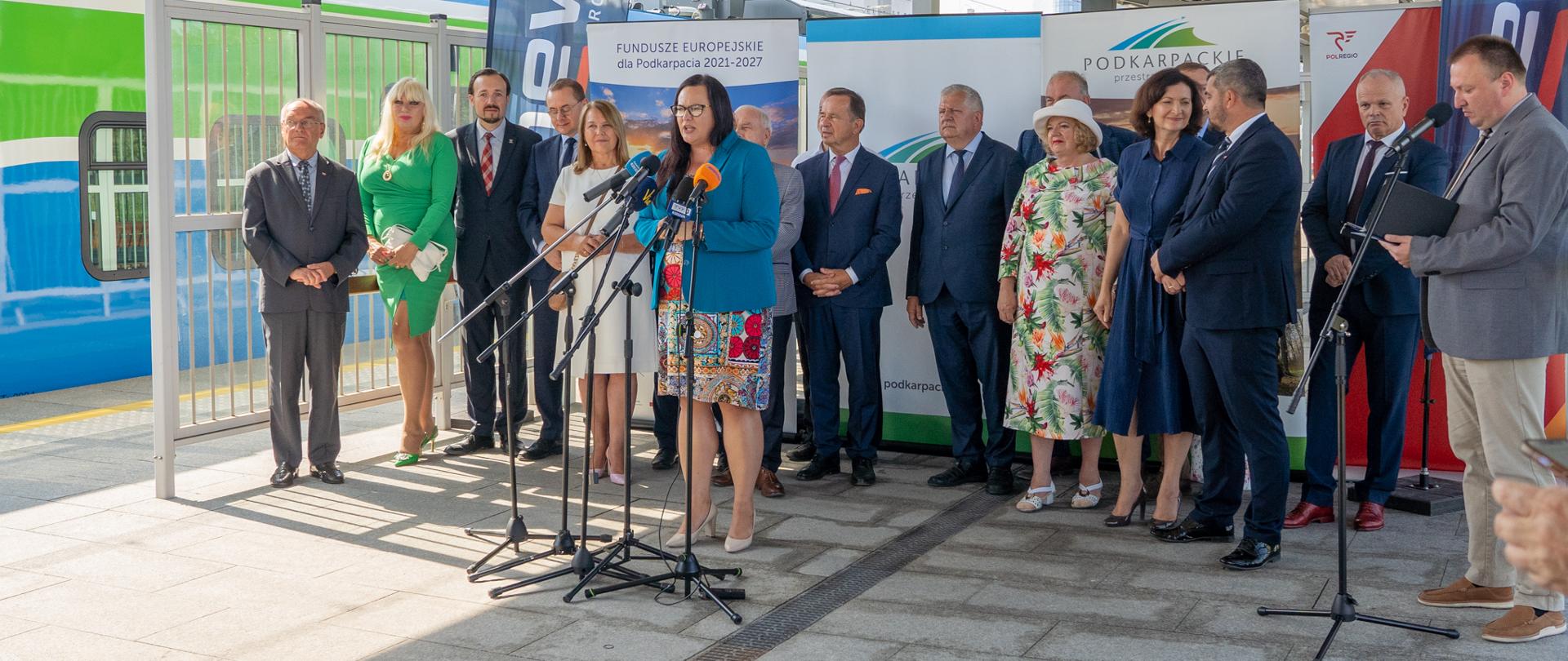 Grupa osób stoi na peronie dworca. Z Przodu przed mikrofonami wiceminister Małgorzata Jarosińska-Jedynak.