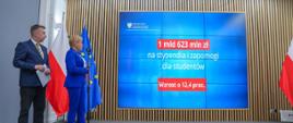 W sali stoją minister Wieczorek i wiceminister Mrówczyńska, patrzą na wbudowany w ścianę wielki ekran z napisem 1 mld 623 mln zł na stypendia i zapomogi dla studentów - wzrost o 12,4 proc.