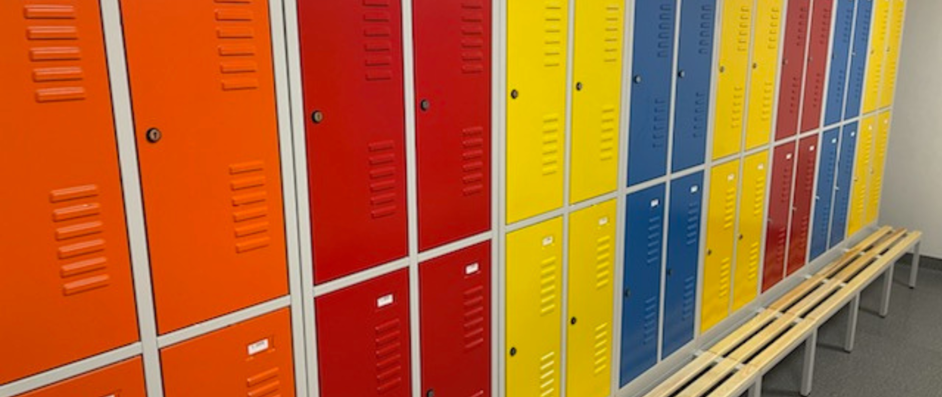 Na zdjęciu kolorowe ustawione w szeregach szafki szatniowe dla uczniów. Szafki są w kolorach: pomarańczowe, czerwone, żółte i niebieskie. Pod szafkami ławeczka z drewnianymi listewkami na siedzisku.