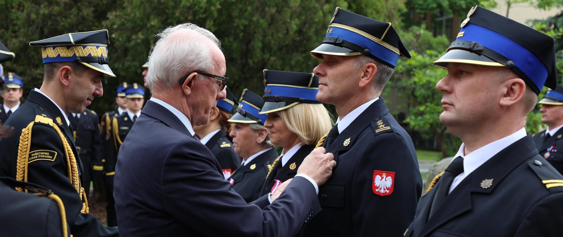 Wiceminister SWiA wraz z komendantem głównym PSP wręczają odznaczenia strażakom PSP, którzy stoją w mundurach galowych na placu 