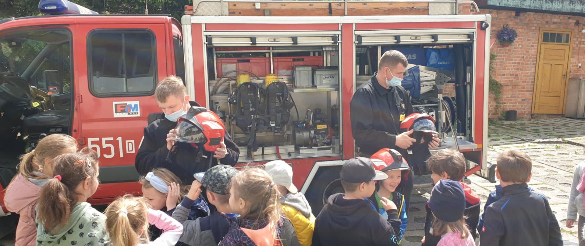 Na zdjęciu widać dwóch strażaków w maseczkach ochronnych oraz grupę przedszkolaków. W tle ustawiony jest samochód strażacki ze sprzętem ratowniczym. Strażacy zakładają dzieciom hełmy.