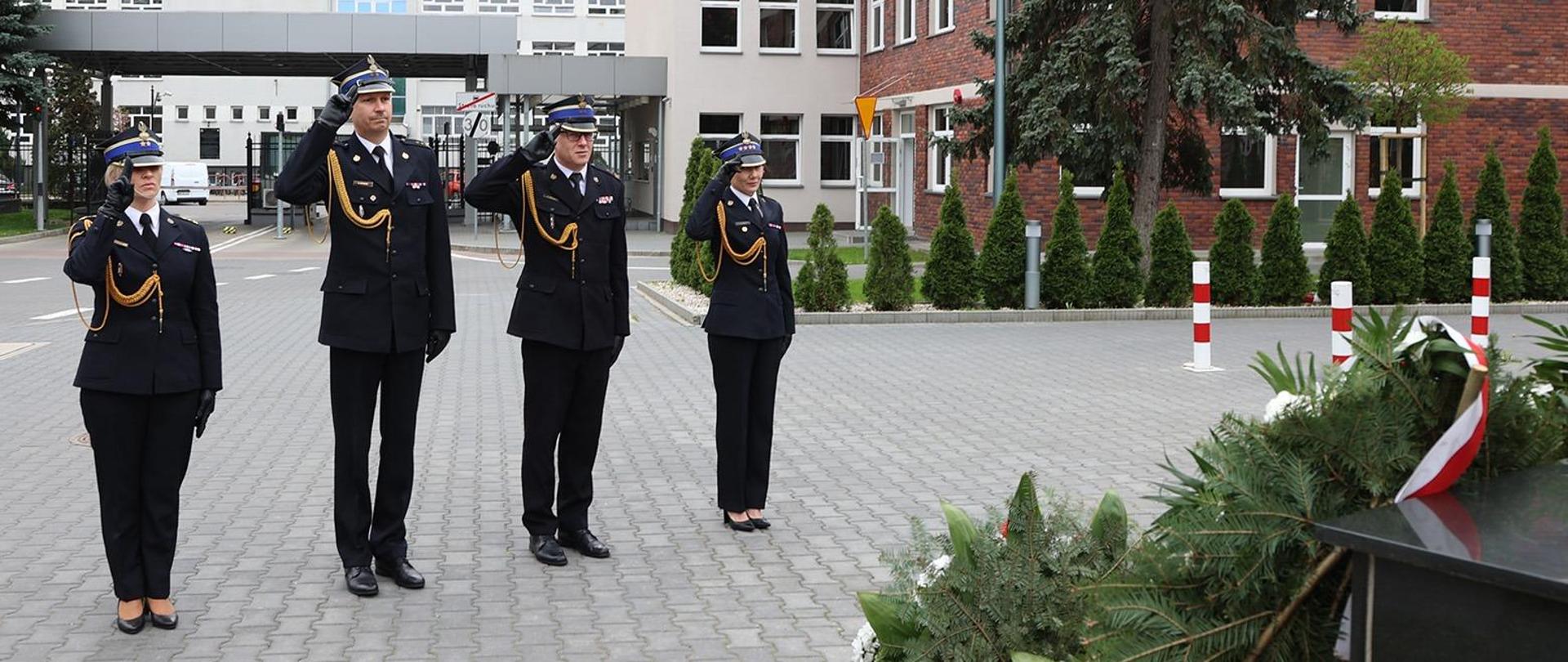 Dwie kobiety i dwóch mężczyzn w strażackich mundurach galowych w momencie salutowania. W tle budynki i brama wjazdowa.