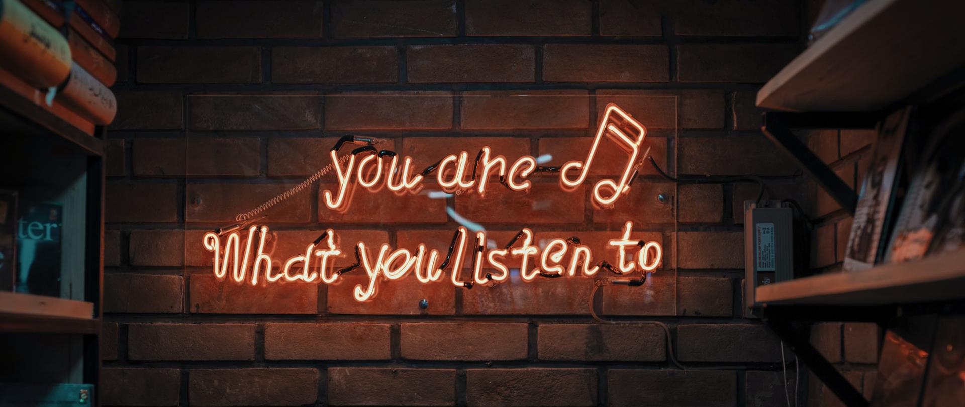 zdjęcie przedstawiające neon o treści "you are what you listen to" zawieszony na ścianie z cegieł, po obu stronach neonu znajdują się półki z książkami