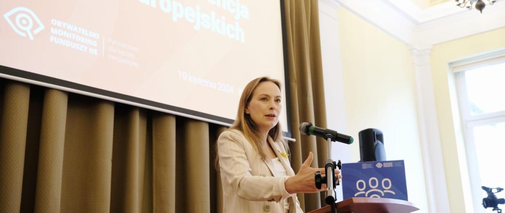 Minister Katarzyna Pełczyńska-Nałęcz stoi przy pulpicie z prawą ręką wyciągniętą do przodu i mówi do mikrofonu.