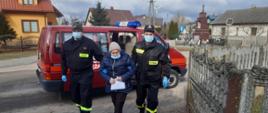Dwóch strażaków OSP prowadzi starszą kobietą z samochodu osobowego strażackiego do domu.