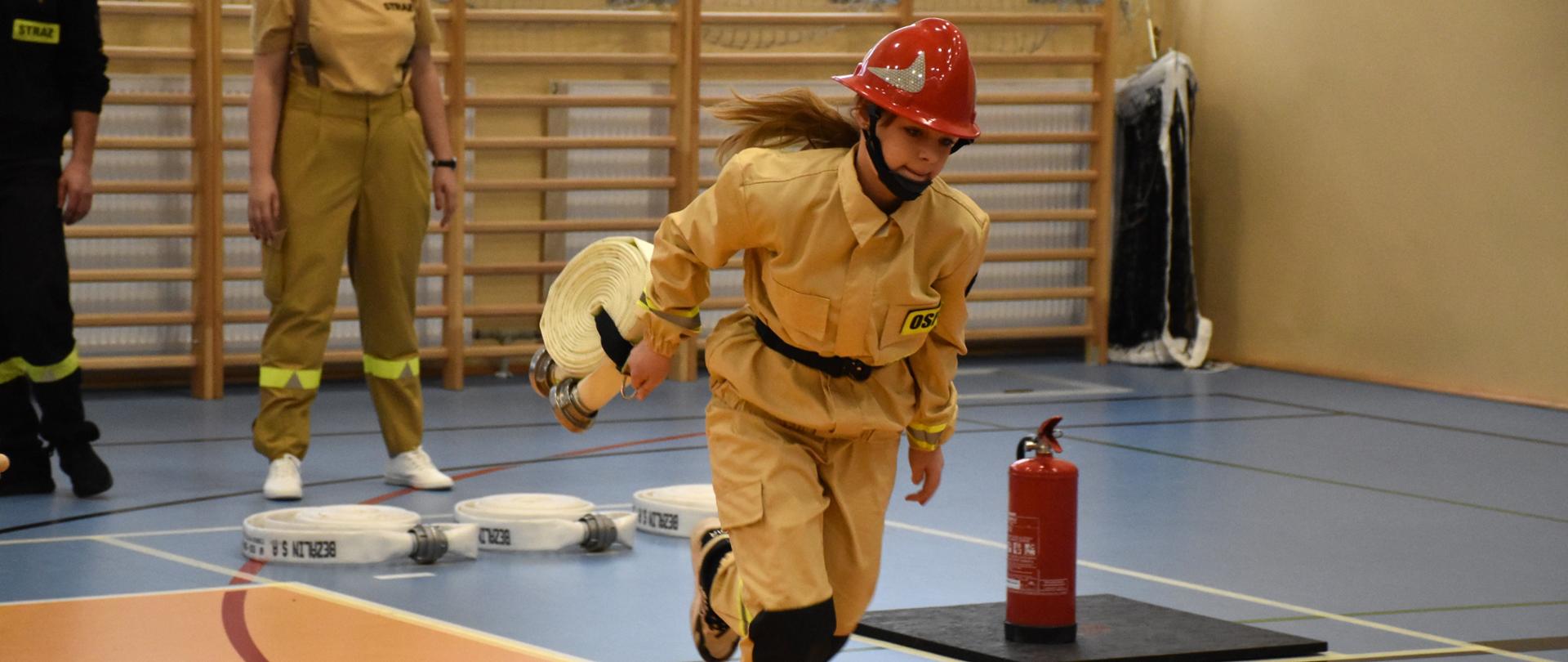 Dziewczynka z Młodzieżowej Drużyny Pożarniczej podczas konkurencji, biegnie po torze trzymając w lewym ręku odcinek węża strażackiego z noszakiem, w tle obserwujący wykonanie zadania druhna i sędzia