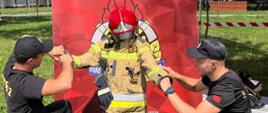 Dwóch strażaków pomaga ubrać się chłopcu w bluzę umundurowania specjalnego. Chłopiec na głowie ma hełm strażacki. 