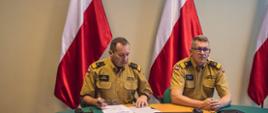Dwaj Zastępcy Komendantów Głównych PSP siedzący przy stole na tle flag narodowych oraz godła Polskiego.