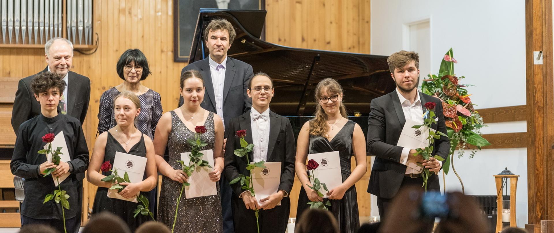 Zdjęcie przedstawia Laureatów i Jurorów XVIII Festiwalu Pianistycznego Chopinowskie Interpretacje Młodych, stojących na scenie w strojach galowych z dyplomami i kwiatami. Jurorzy stoją w drugim rzędzie, widać publiczność, w tle fortepian i portret I. J, Paderewskiego.