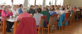 Spotkanie z seniorami. Sala w jednej z rawickich restauracji. Po obu stronach stołu siedzą uczestnicy spotkania, seniorki i seniorzy z osiedla Sienkiewicza w Rawiczu (ponad 50 osób). 