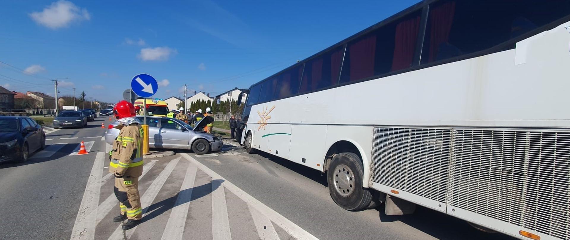 Zdjęcie przedstawia po prawej stronie stojący na pasie jezdni uszkodzony autobus turystyczny, a przed nim, ustawiony do niego prostopadle samochód osobowy. Na drodze stoi strażak, a w głębi widać ambulans.