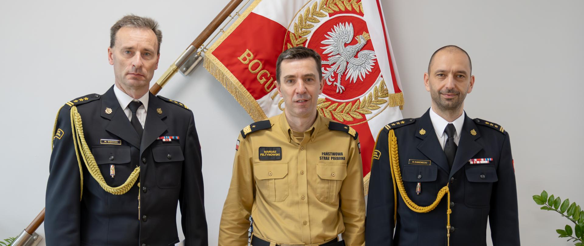 Trzech oficerów Państwowej Straży Pożarnej pozuje do pamiątkowej fotografii. Na zdjęciu stoi komendant główny PSP, warmińsko-mazurski komendant wojewódzki oraz pełniący obowiązki zastępcy warmińsko-mazurskiego komendanta wojewódzkiego.