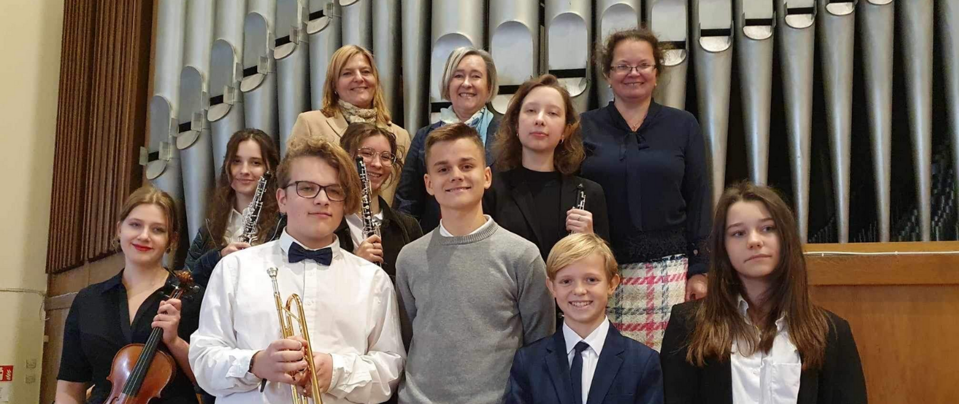 Na zdjęciu grupa uśmiechniętej młodzieży. Wszyscy odświętnie ubrani. Niektórzy trzymają instrumenty muzyczne: skrzypce, trąbkę, klarnet. W tle piszczałki organowe.