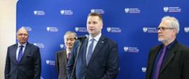 Minister Czarnek stoi przed niebieską ścianka i mówi do mikrofonu na stojaku, obok niego trzech mężczyzn w garniturach.