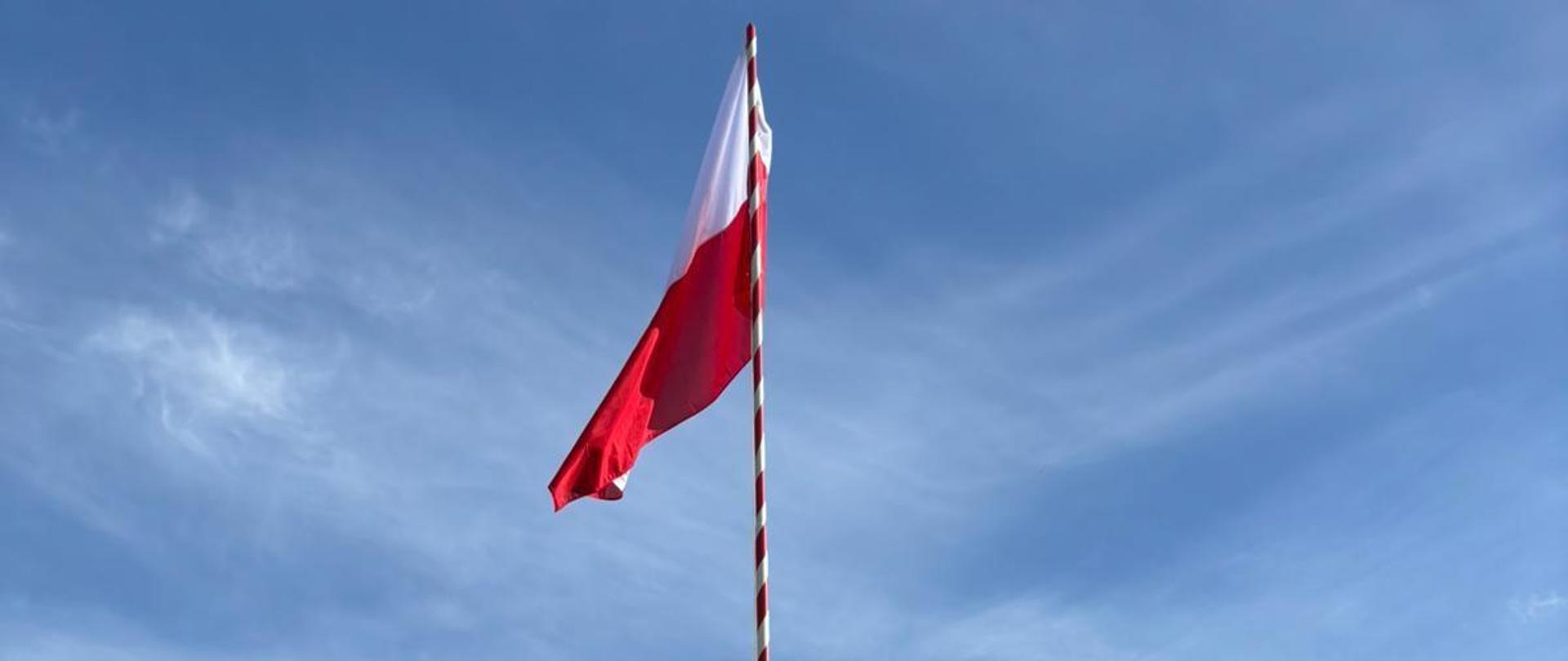 Biało-czerwona flaga Rzeczypospolitej Polskiej powiewa na maszcie, na tle błękitnego nieba.